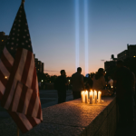 An American flag at dusk with two beams of light rising into the sky and lit candles on a stone ledge, silhouetted figures gathered nearby in a September 11th memorial tribute scene